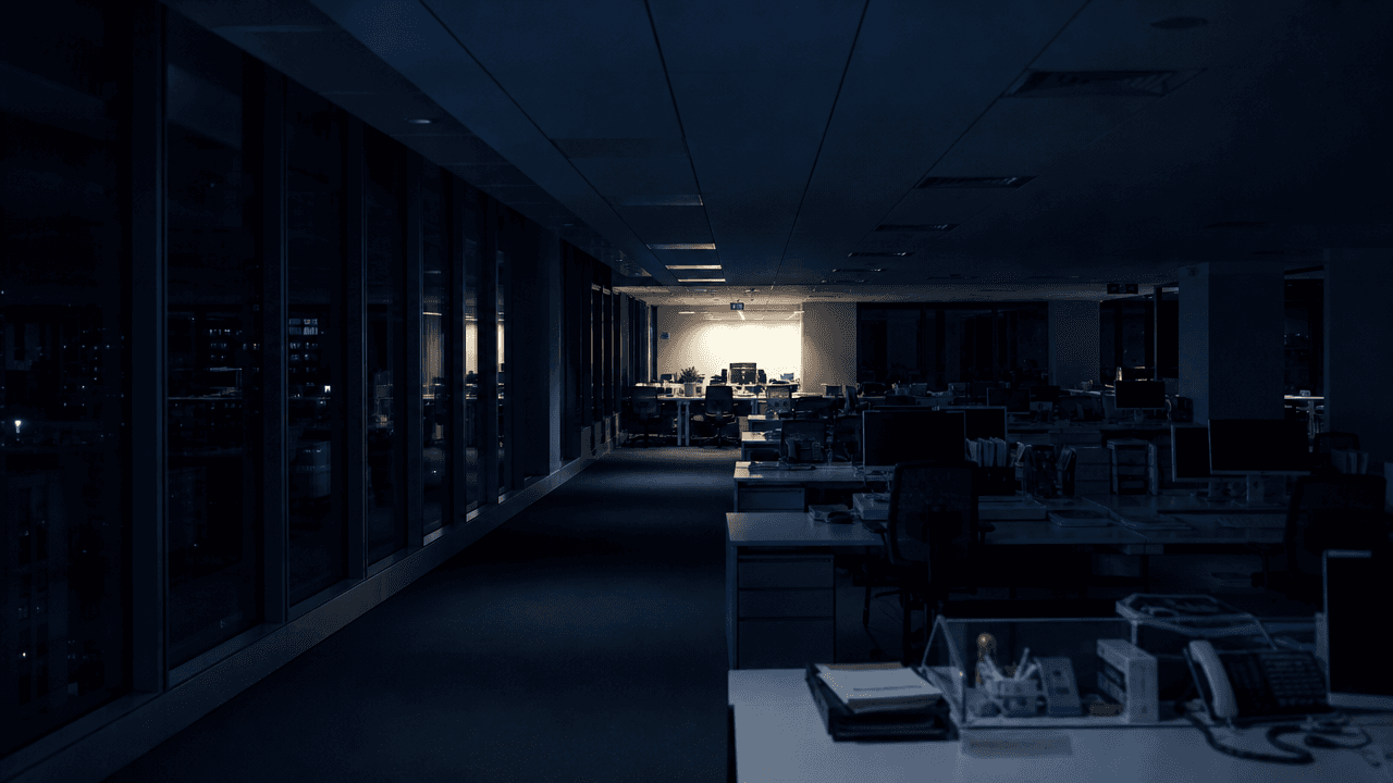 Plateau de bureaux plongé dans l'obscurité, une seule zone éclairée au fond, pendant une vacation de nuit